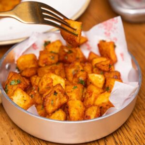 Close-up of crispy spicy potato cubes in a bowl with meal setting on a wooden table.