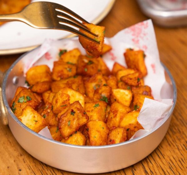 Close-up of crispy spicy potato cubes in a bowl with meal setting on a wooden table.